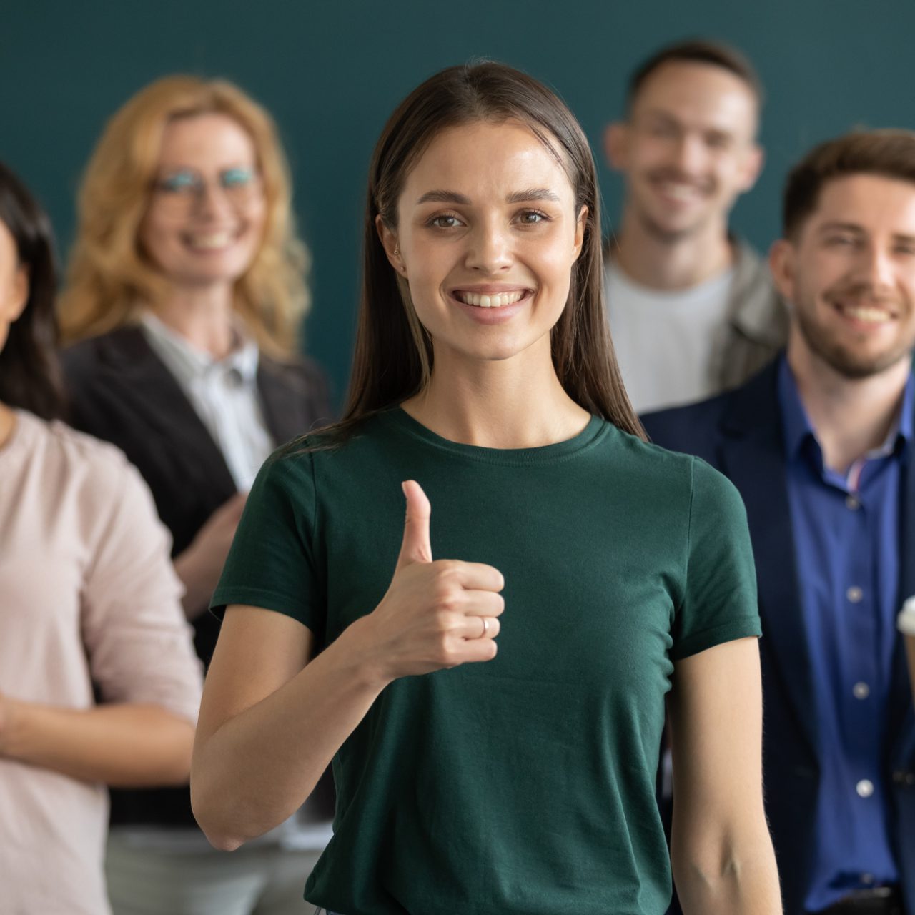 Happy young female business leader making like thumb up, diverse team smiling in background. Satisfied customer, employee with multiethnic group behind giving positive feedback. Head shot portrait