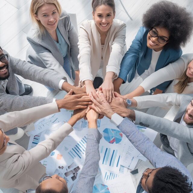 A group of diverse business professionals sitting in a circle, putting their hands together in a team gesture, top-down view, teamwork and unity, charts and documents on the floor, modern office, collaboration, positive energy, high resolution, sharp focus, professional commercial stock photo style --no watermark, no text, no logo, no brand --ar 16:9 --v 7 Job ID: 64ac25e2-9085-420a-8aec-fac7f51ae802